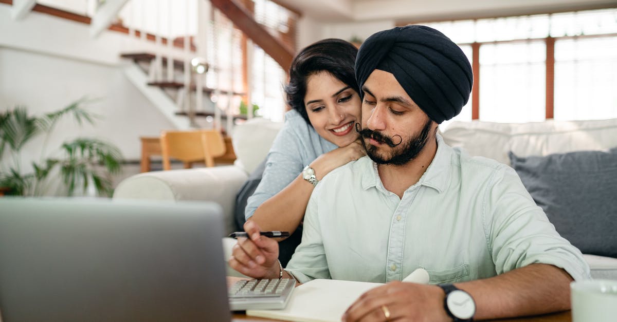 Account and downloading help - Cheerful young Indian woman cuddling and supporting serious husband working at home with laptop and counting on calculator Account and downloading help - Cheerful young Indian woman cuddling and supporting serious husband working at home with laptop and counting on calculator