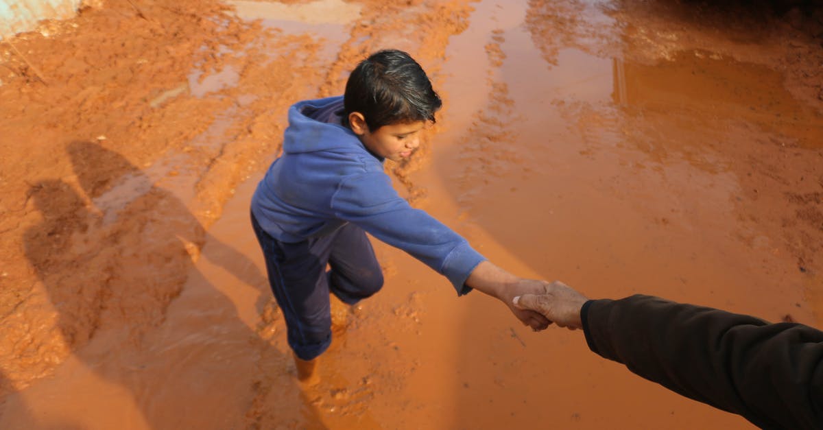 Am I stuck in a boy band? - High angle of crop person holding hands with ethnic boy stuck in dirty puddle in poor village