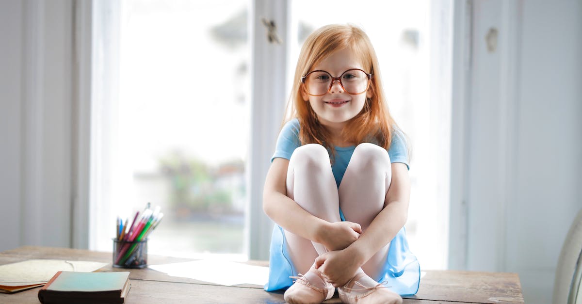 Anywhere to sit inside the Earth Temple? - Joyful red haired schoolgirl in blue dress and ballet shoes smiling at camera while sitting on rustic wooden table hugging knees beside school supplies against big window at home