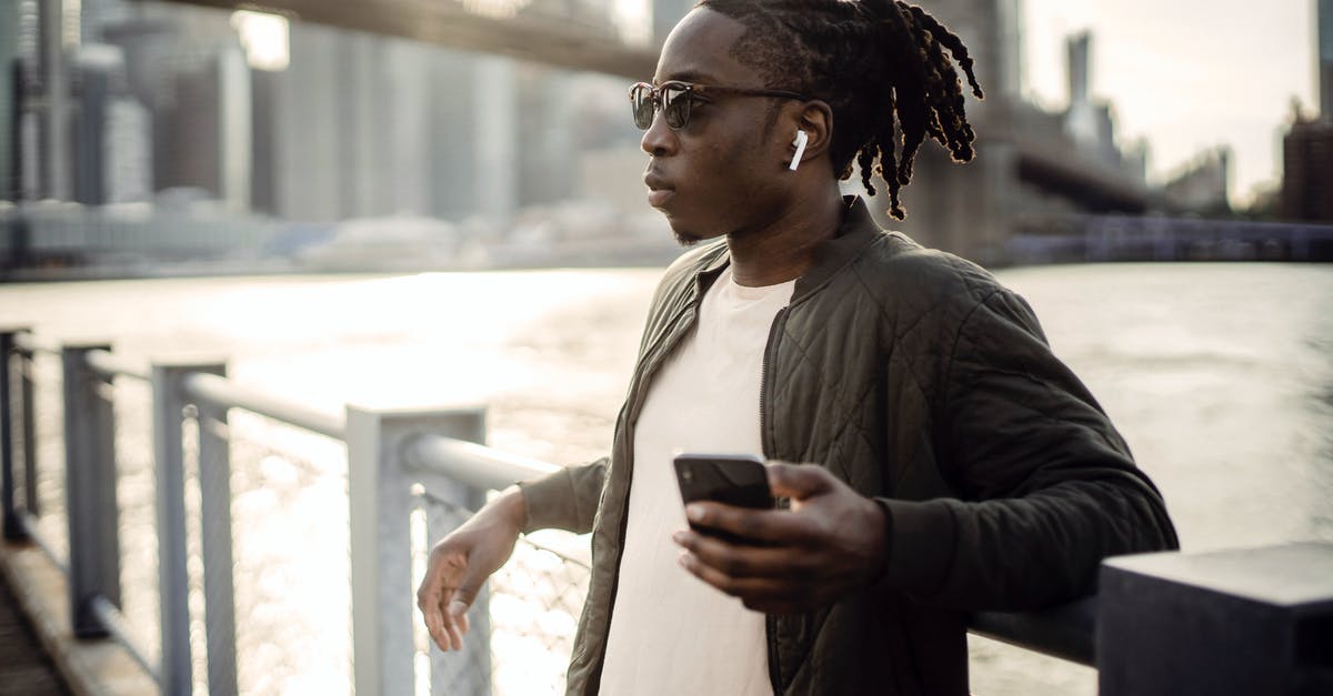 Batman Arkham Series thinks I have surround sound - Thoughtful masculine African American guy in sunglasses and true wireless earphones listening to music from cellphone while standing leaned on bridge fence passing over city river Batman Arkham Series thinks I have surround sound - Thoughtful masculine African American guy in sunglasses and true wireless earphones listening to music from cellphone while standing leaned on bridge fence passing over city river