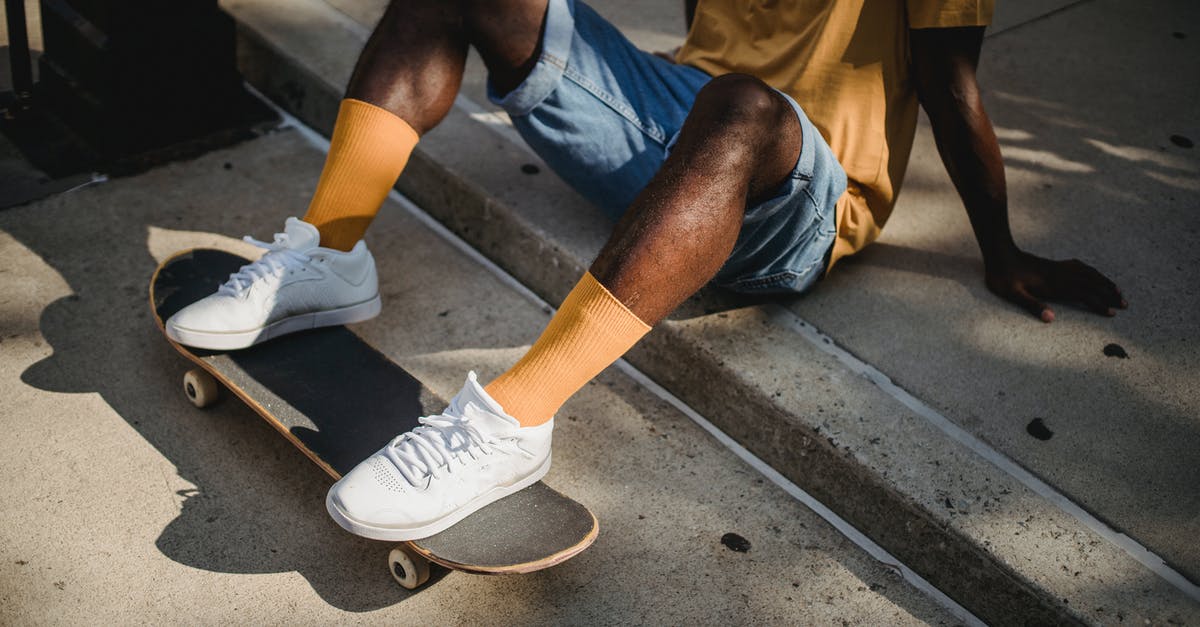 Can't do a mob invisible trick - Crop anonymous African American male skater in t shirt and denim shorts sitting on sidewalk with feet on skateboard