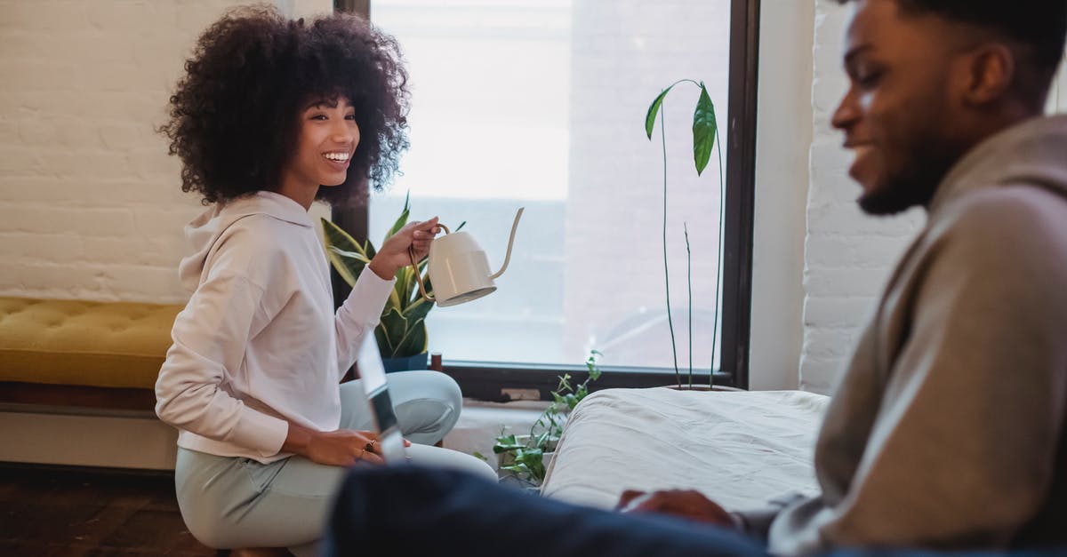 Can Genji deflect when using Dragonblade? - Smiling African American woman in casual clothes squatting down and watering plants while spending time with boyfriend browsing laptop at home in daytime
