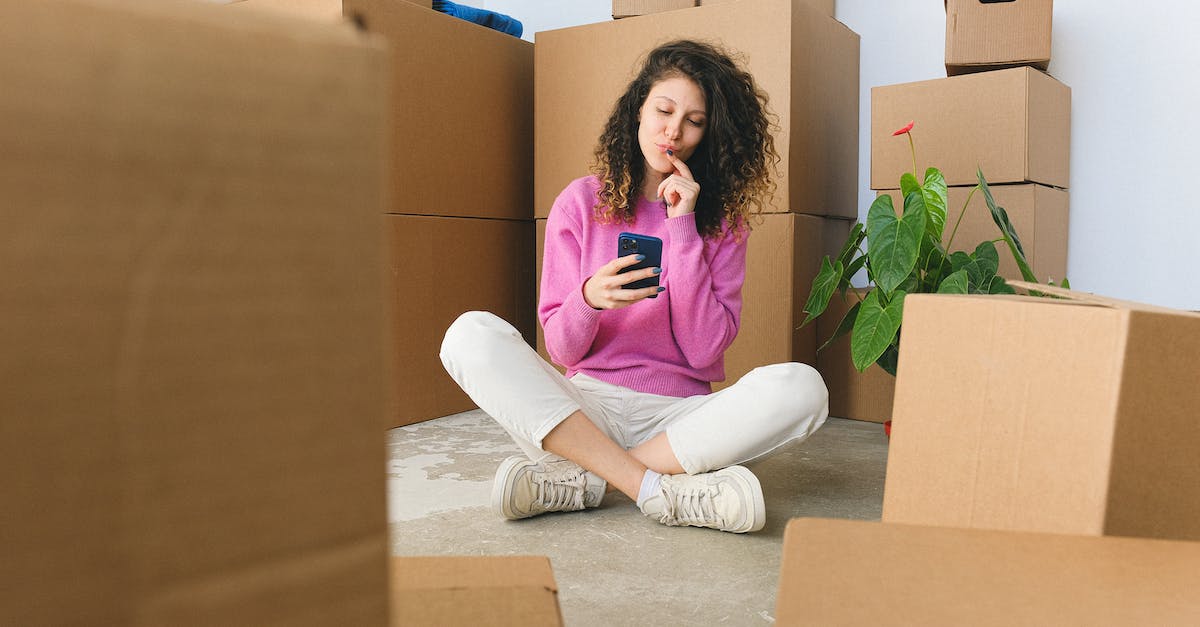 Can I copy the content of my message box (using backup utility)? - Full body of positive thoughtful young female millennial in casual outfit touching lips and using smartphone while sitting on floor with crossed legs near carton boxes after relocation