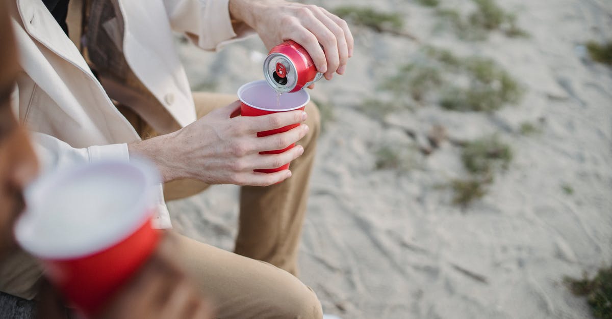 Can I directly battle my friends? - High angle of crop anonymous male pouring refreshing drink into red cup while sitting on sandy shore with black friend