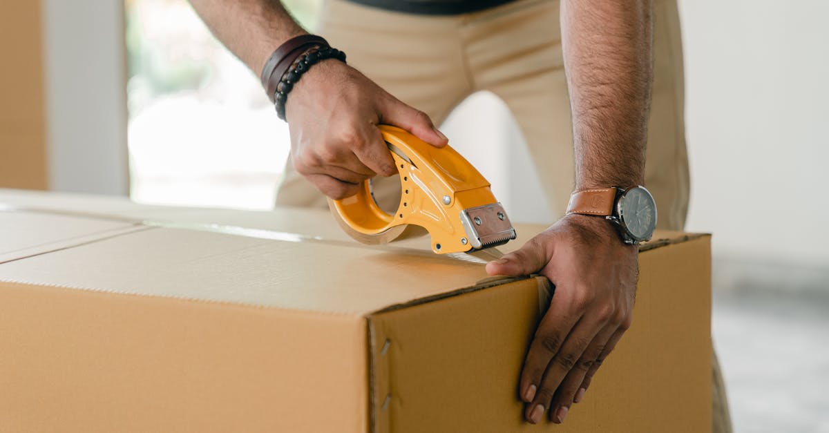 Can I move a house a few inches? - Crop faceless young male with wristwatch using adhesive tape while preparing cardboard box for transportation