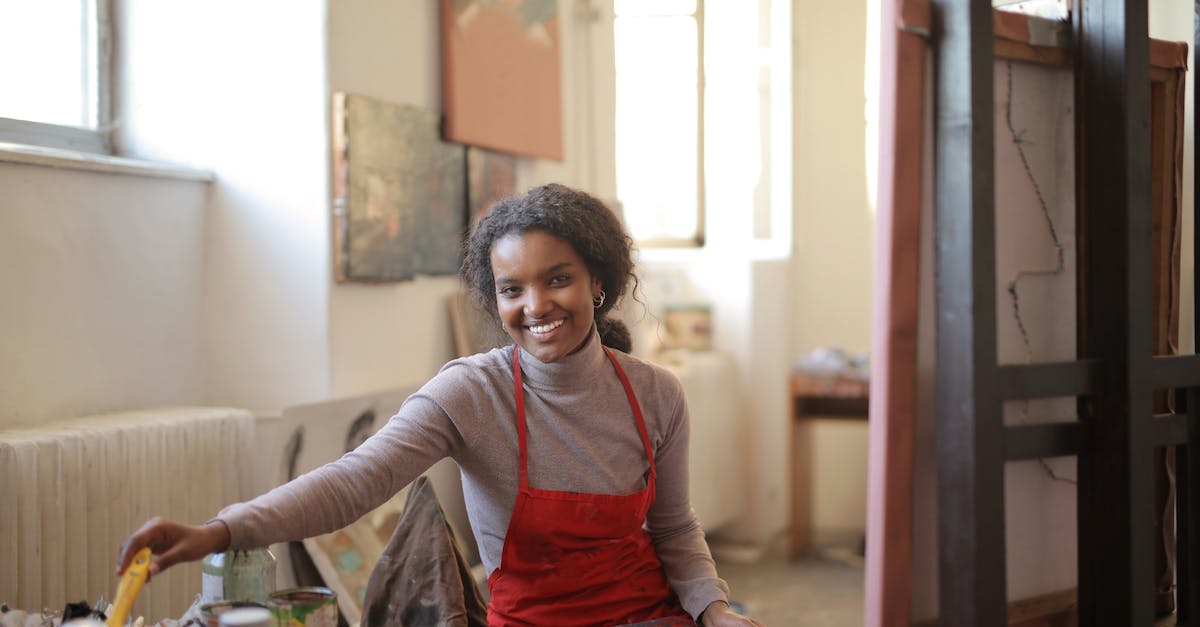 Can I put black paint on a cow mangler? - Cheerful smiling African American female in apron sitting on chair in front of easel near table with various tools for painting in creative workshop