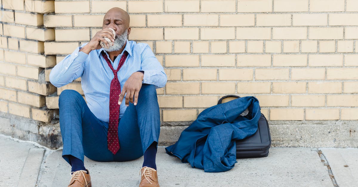 Can you dodge the blue shell? - Full body of mature African American bearded businessman in blue trousers and light shirt with maroon tie sitting on ground at brick wall and drinking beverage from tin can