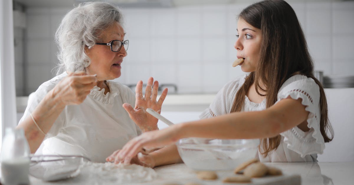 Changing Xbox settings from Adult to child or teen - Calm senior woman and teenage girl in casual clothes looking at each other and talking while eating cookies and cooking pastry in contemporary kitchen at home