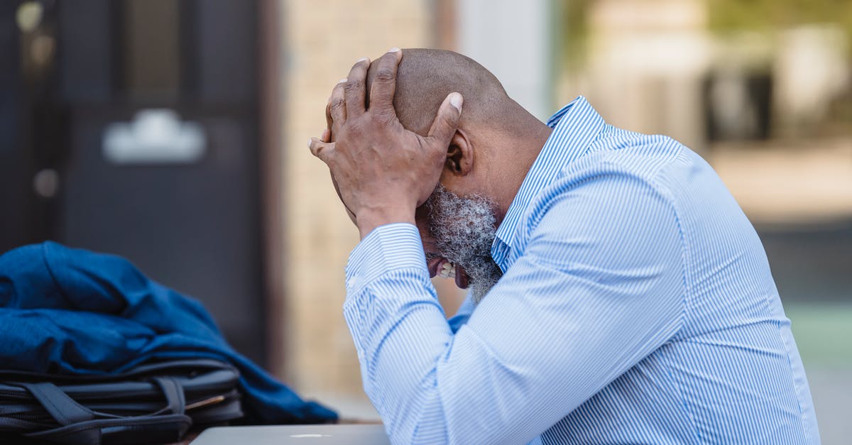 Cheating expert bosses - Side view of pensive middle aged African American male entrepreneur wearing blue shirt grabbing head with hands while sitting at table with closed laptop in veranda