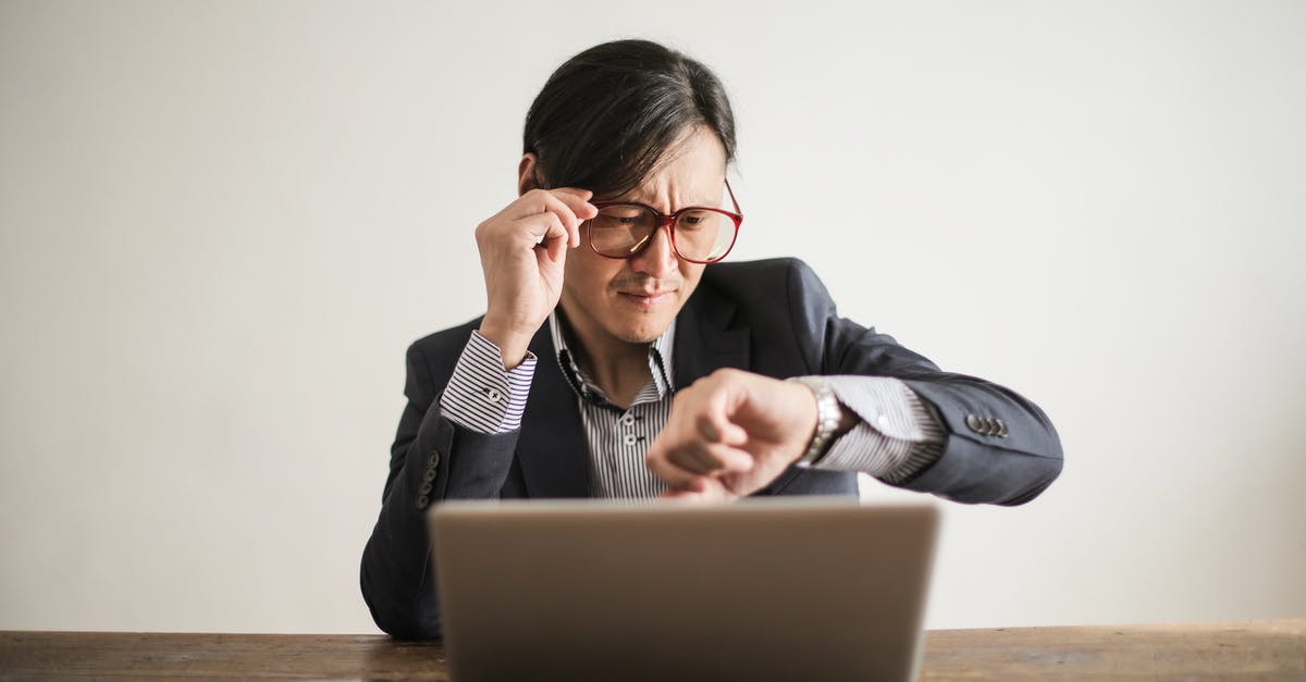 Correlation between Rushing and Time Remaining on a Task? - Young frowning man in suit and glasses looking at wristwatch while waiting for appointment sitting at desk with laptop Correlation between Rushing and Time Remaining on a Task? - Young frowning man in suit and glasses looking at wristwatch while waiting for appointment sitting at desk with laptop