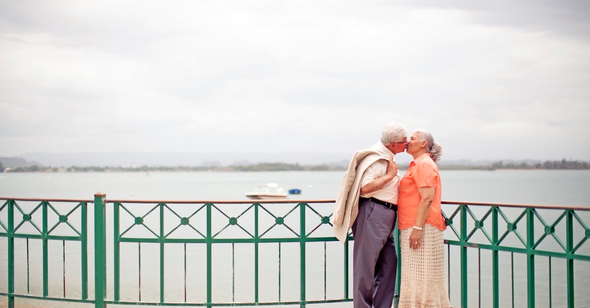Dating in Style Savvy: Trendsetters? - Side view full body romantic senior couple wearing trendy outfits standing on waterfront promenade and kissing gently on overcast weather Dating in Style Savvy: Trendsetters? - Side view full body romantic senior couple wearing trendy outfits standing on waterfront promenade and kissing gently on overcast weather