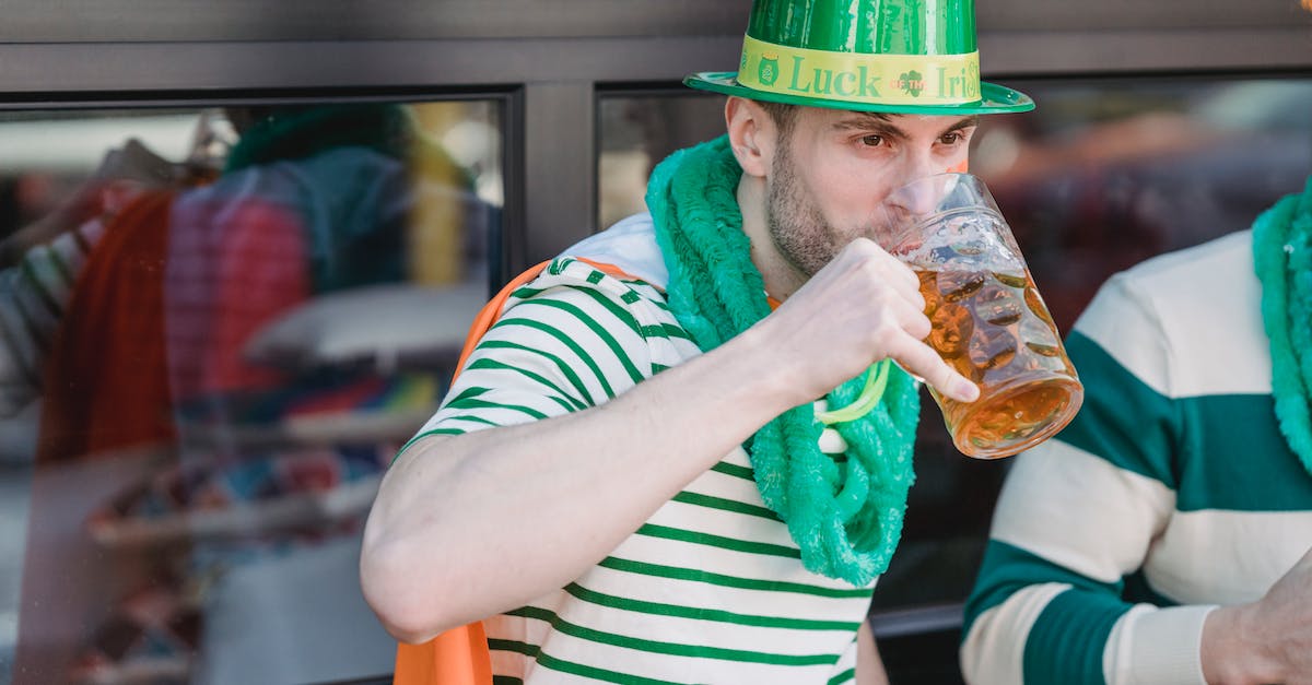 Do Luck-J and Mug synergise? - Young unshaven male millennial in casual clothes and green leprechaun hat drinking mug of cold beer and looking away during celebration pf St Patricks Day with crop anonymous friend