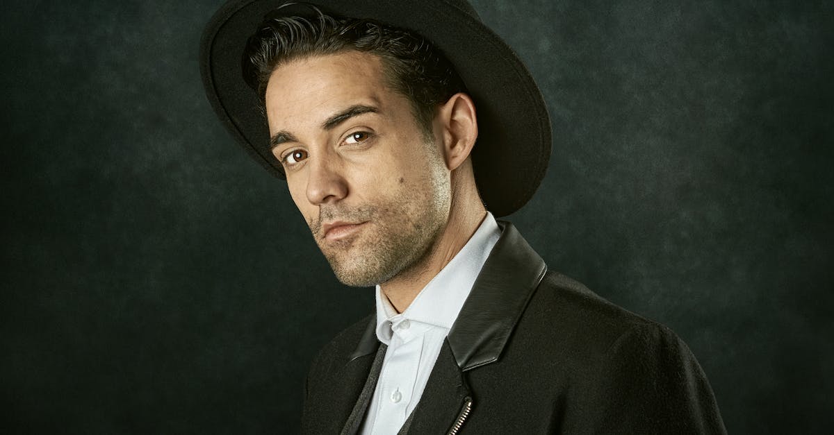 Do most hats only make you look fancy? - Portrait of stylish positive young Hispanic male wearing elegant black suit and hat standing against black background in studio and looking at camera Do most hats only make you look fancy? - Portrait of stylish positive young Hispanic male wearing elegant black suit and hat standing against black background in studio and looking at camera