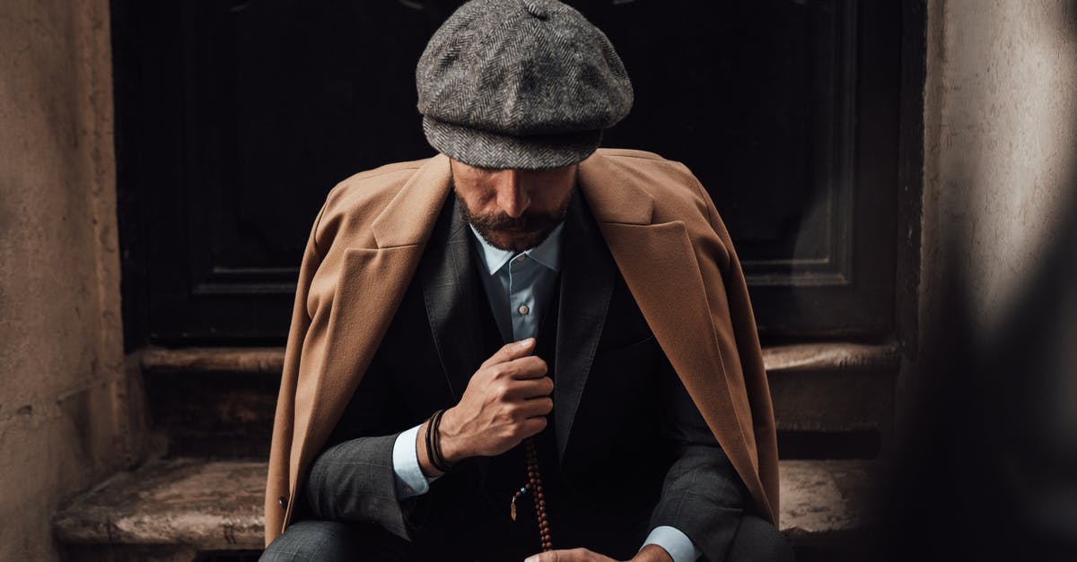 Do most hats only make you look fancy? - Pensive stylish male in posh suit coat and hat sitting with beads on shabby doorway and looking down in thoughts Do most hats only make you look fancy? - Pensive stylish male in posh suit coat and hat sitting with beads on shabby doorway and looking down in thoughts