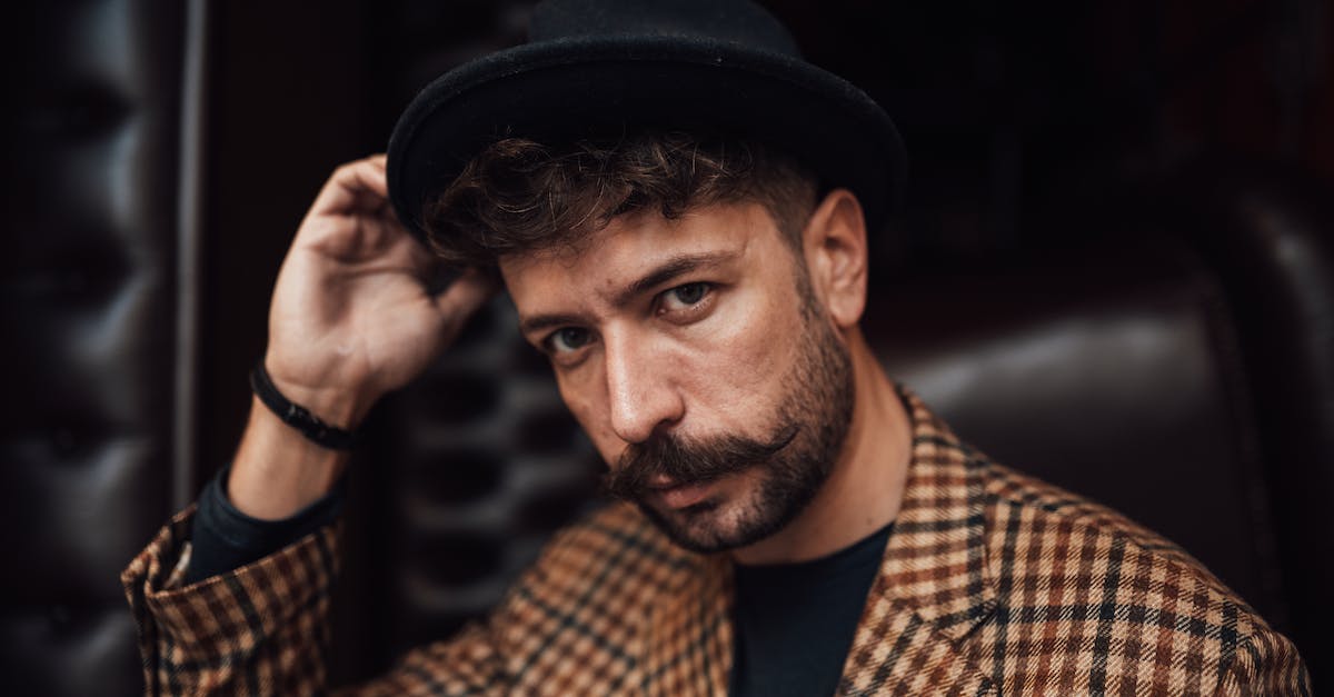 Do most hats only make you look fancy? - Unemotional adult male with beard and mustache wearing classy suit touching hat brim and looking at camera while sitting on comfy leather couch Do most hats only make you look fancy? - Unemotional adult male with beard and mustache wearing classy suit touching hat brim and looking at camera while sitting on comfy leather couch