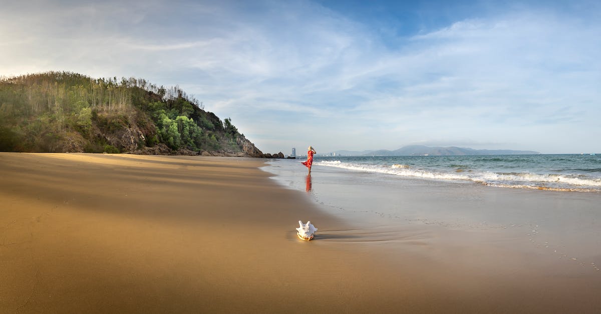 Dodging red shell - Distant female wearing red maxi dress and strolling on wet sandy seacoast against lush hilly cliff on sunny summer day