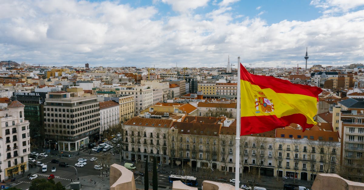 Does National visistor center affect all cities? - Drone view of Spanish city with aged buildings and national flag under cloudy blue sky Does National visistor center affect all cities? - Drone view of Spanish city with aged buildings and national flag under cloudy blue sky