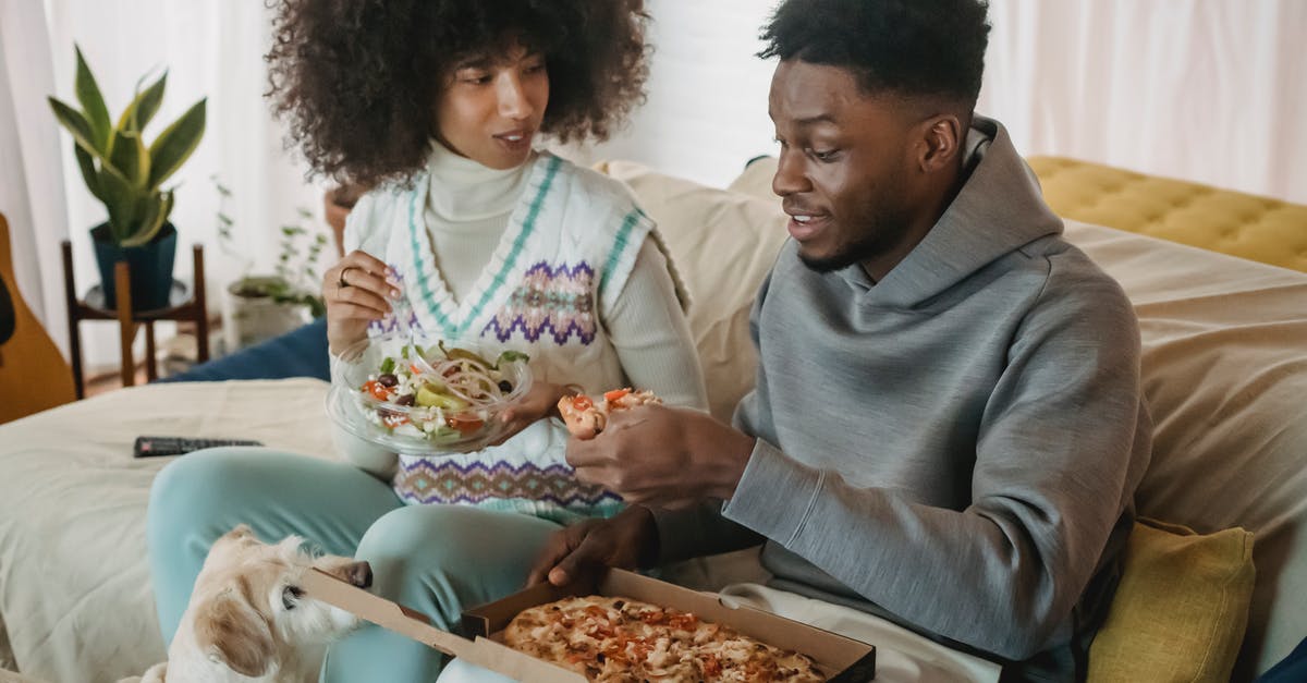 Dog Salad RNG Debunk - Positive African American couple eating salad and pizza while sitting on couch near cute dog in cozy living room at home