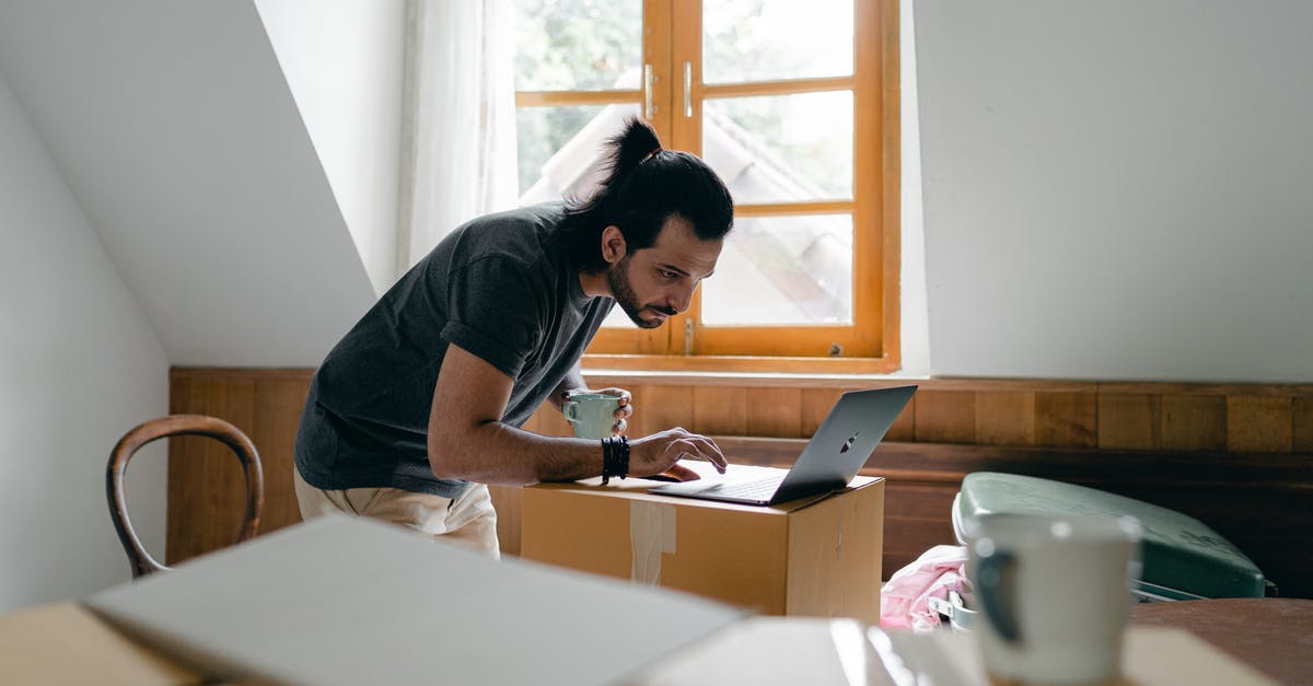 Enigma data pack in the Royal Hotel's ballroom is inaccessible - Side view of focused bearded male in casual outfit with cup of hot drink browsing laptop while packing stuff into carton boxes Enigma data pack in the Royal Hotel's ballroom is inaccessible - Side view of focused bearded male in casual outfit with cup of hot drink browsing laptop while packing stuff into carton boxes