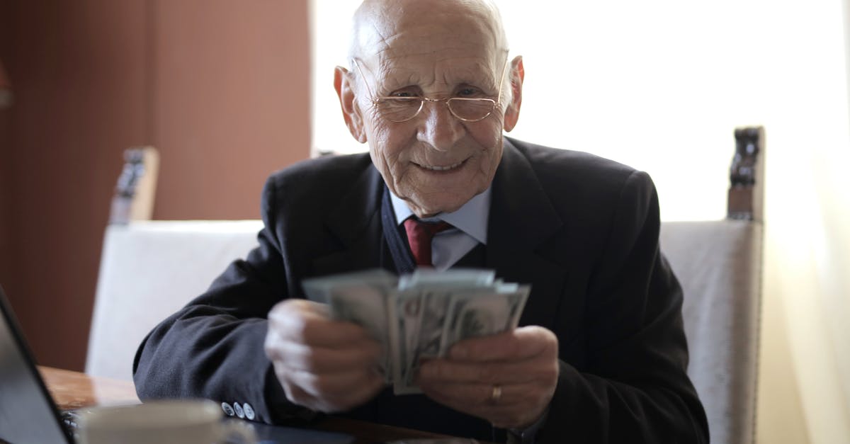Fifa career and stadium capacity - Positive senior businessman in formal suit and eyeglasses counting money bills while sitting at wooden table with cup of beverage and near opened laptop