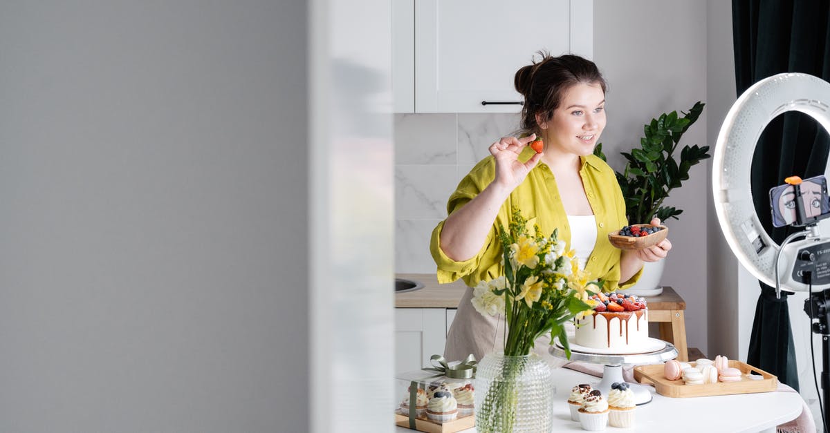 Fortnite stuck before showing intro video - Delighted young female influencer in casual clothes smiling and demonstrating fresh berries while decorating appetizing cake during recording vlog on smartphone in kitchen