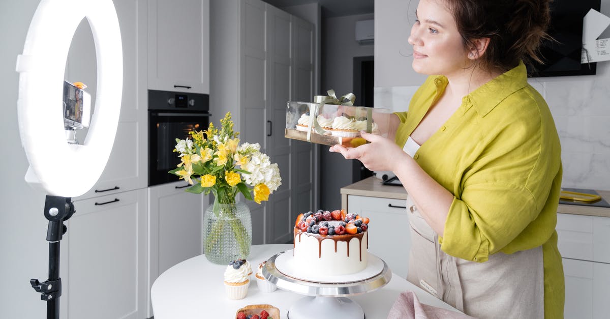 Fortnite stuck before showing intro video - Side view of positive young female food blogger in casual clothes and apron introducing yummy cakes and smiling while standing in front of ring lamp and recording vlog on smartphone