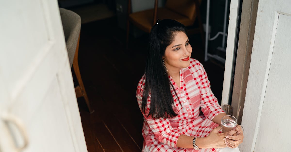 From the water to the door - From above of young Indian lady in casual outfit sitting on floor in doorway with glass of water looking away thoughtfully From the water to the door - From above of young Indian lady in casual outfit sitting on floor in doorway with glass of water looking away thoughtfully