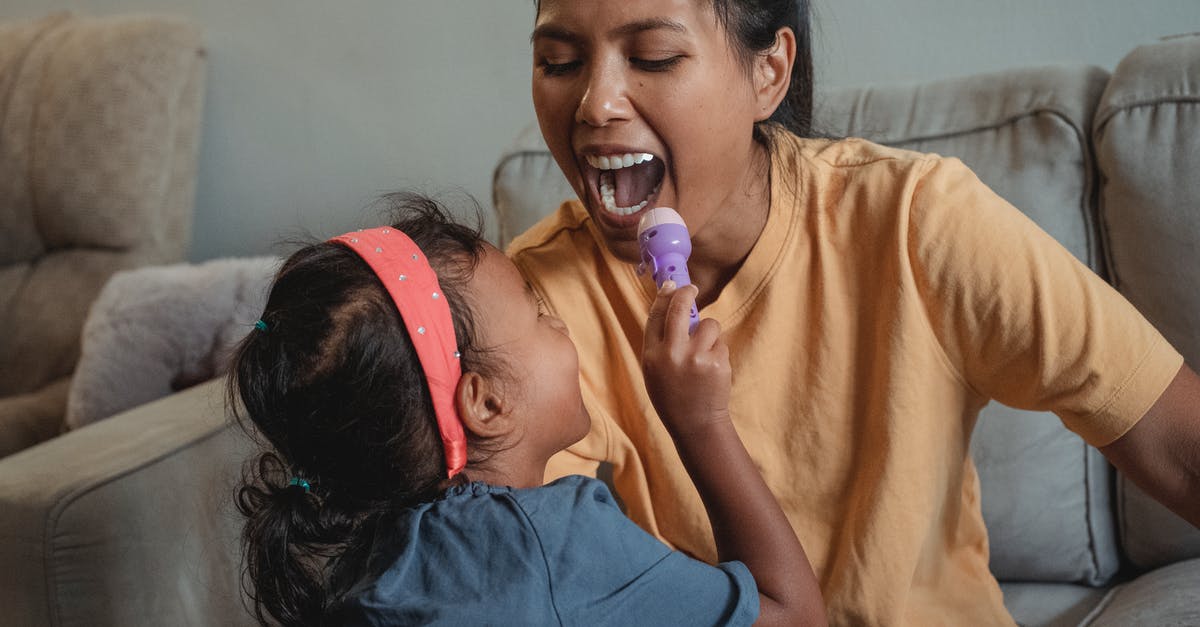 Game won't open? - Cheerful ethnic little girl shining flashlight in mouth of positive mother while doing examination during game on sofa in apartment Game won't open? - Cheerful ethnic little girl shining flashlight in mouth of positive mother while doing examination during game on sofa in apartment