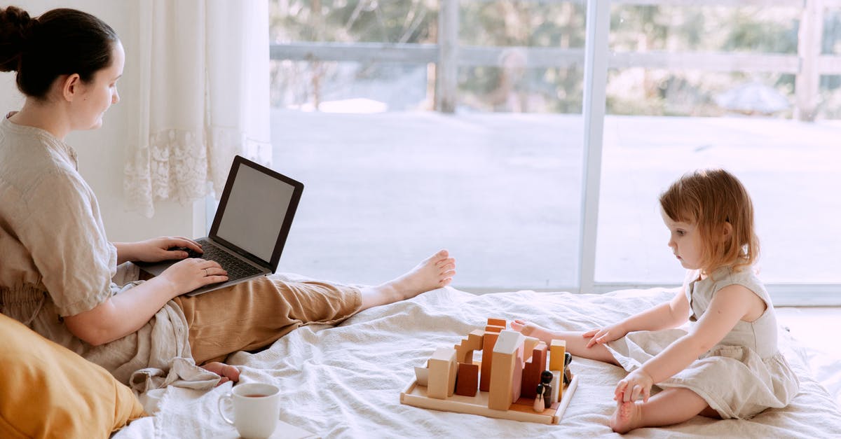 Games not smooth when I play while internet is connected - Side view of casual woman sitting on bed and surfing laptop with blank screen while adorable kid playing with toys in cozy bedroom