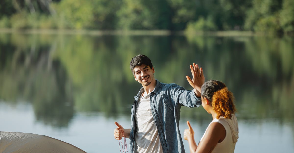 Giving water to strangers - Happy young traveling couple giving each other high five while standing on river bank and putting up tent Giving water to strangers - Happy young traveling couple giving each other high five while standing on river bank and putting up tent