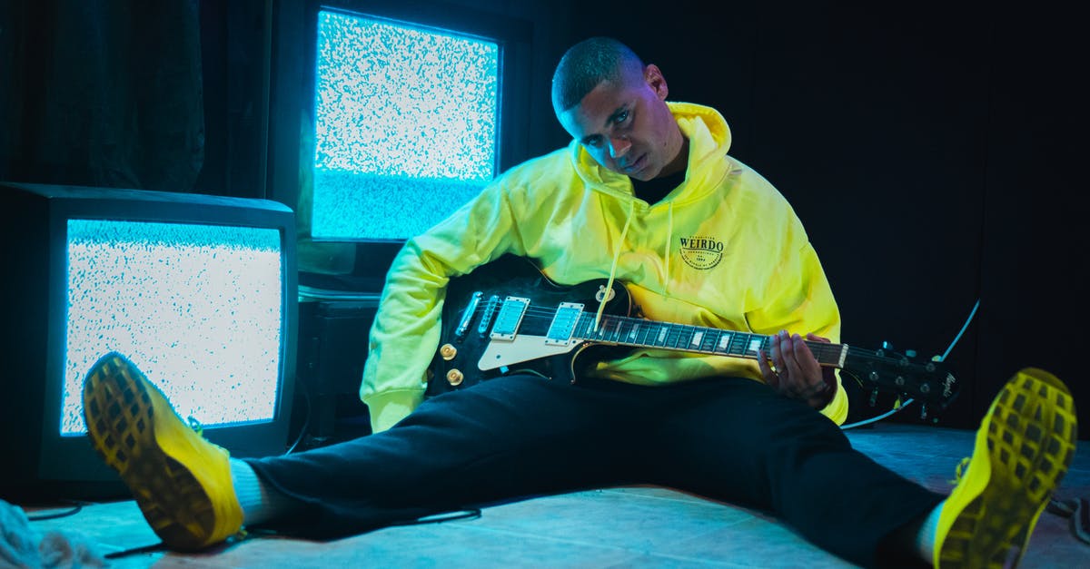 guitar hero 3 freezing at first screen - Young ethnic male guitarist in trendy yellow hoodie and sneakers sitting with guitar on tiled floor near television set with blurry screens and looking at camera