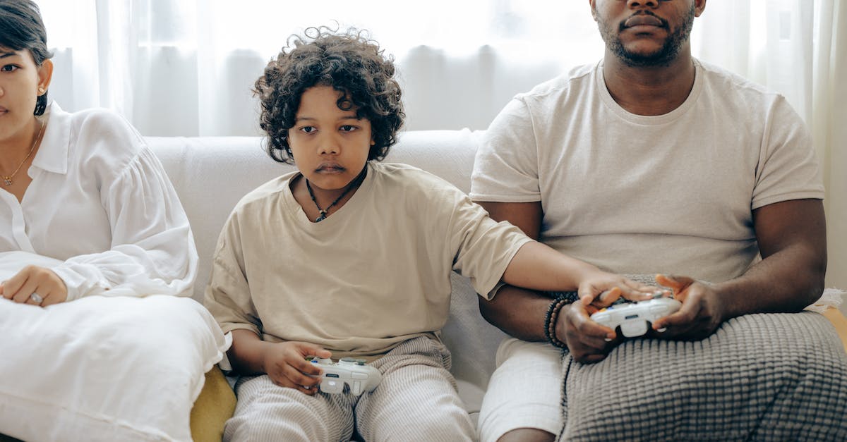 Harrison Jones and its usage - Concentrated child showing usage of gamepad to father while playing on game console and resting on sofa together with family