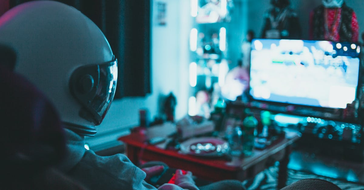 How can I play old console games on a modern TV? - Side view of unrecognizable person in virtual reality helmet sitting on sofa and playing with gamepad in dark room