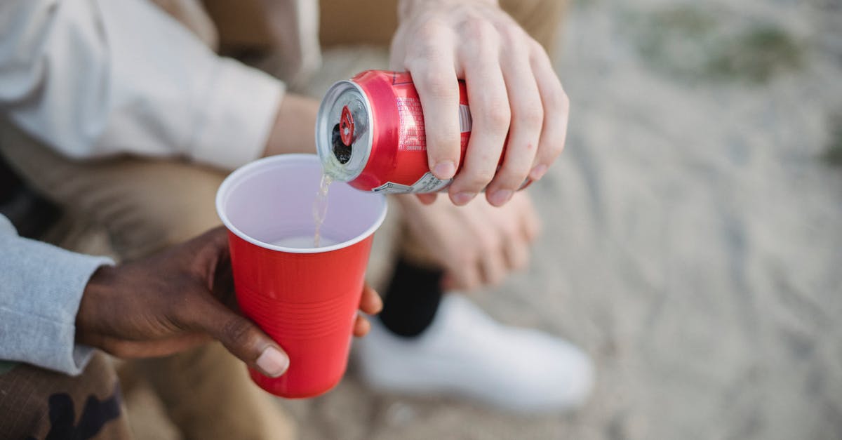 How can I queue spending points? - From above of crop anonymous man pouring fizzy drink from can into red plastic cup of black friend while sitting on sandy ground