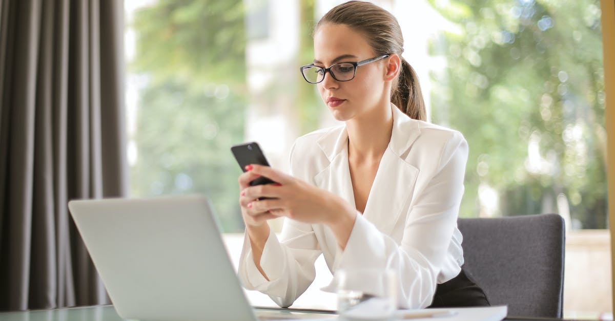 How do I access Looking Glass Knight? - Concentrated young woman in formal clothes and eyeglasses sitting at glass table with laptop while surfing smartphone