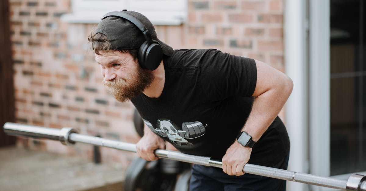 How do I change train direction? - Brutal male with headphones pulling massive metal barbell on blurred background of brick house