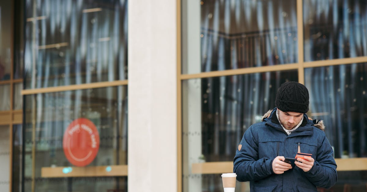How do I check where to go next? - Focused young male in warm outerwear and hat standing near modern building with takeaway coffee and using smartphone while making online shopping with credit card