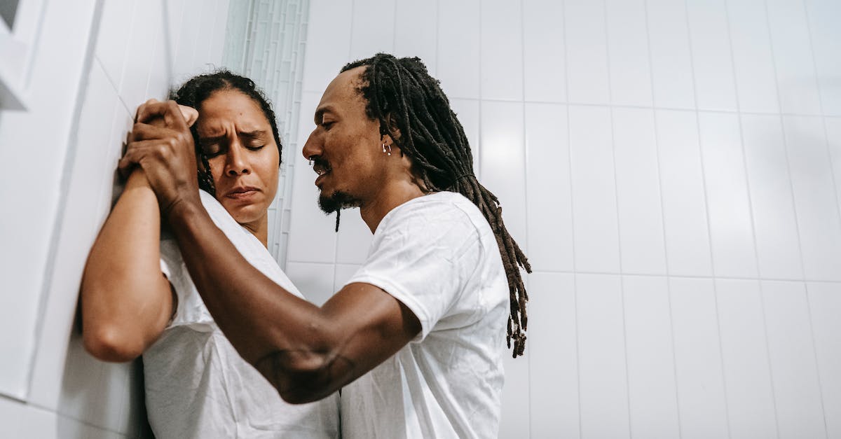 How do I dominate Milligan? - Side view of young black angered man with Afro braids in white t shirt pushing sad wife with closed eyes against tiled wall during conflict in bathroom