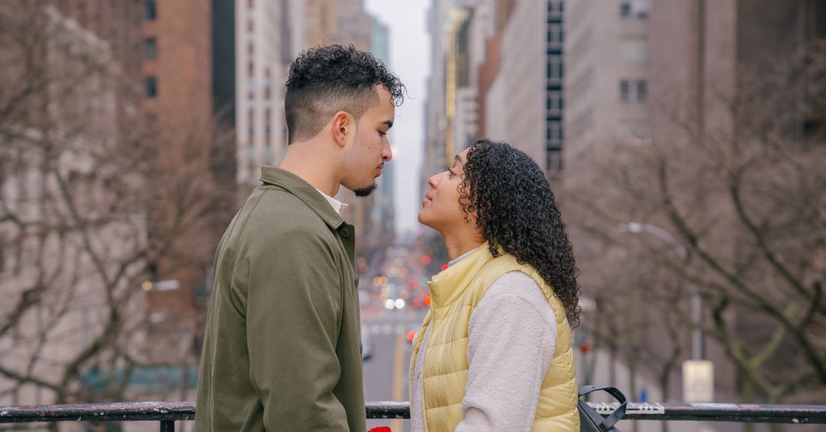 How do I get to the other side of this bridge? - Side view of romantic Hispanic couple in casual clothes looking at each other while standing on bridge near metal fence against city street in daytime