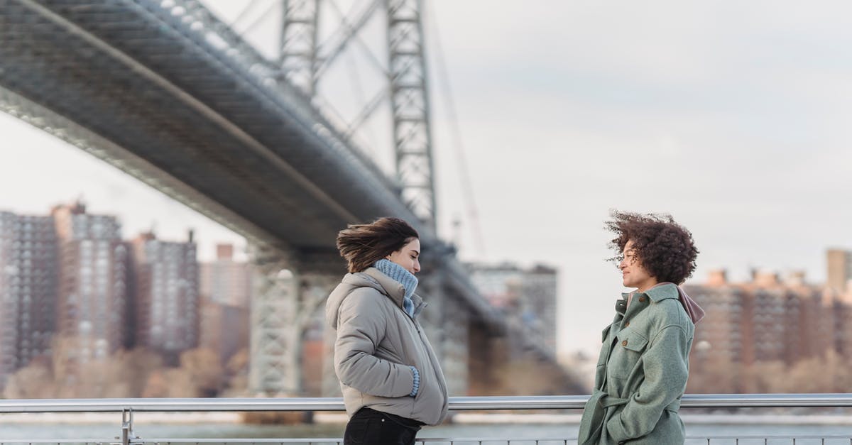 How do I get to the other side of this bridge? - Side view young female friends in warm clothes having conversation and standing face to face on New York City embankment on cold winter day