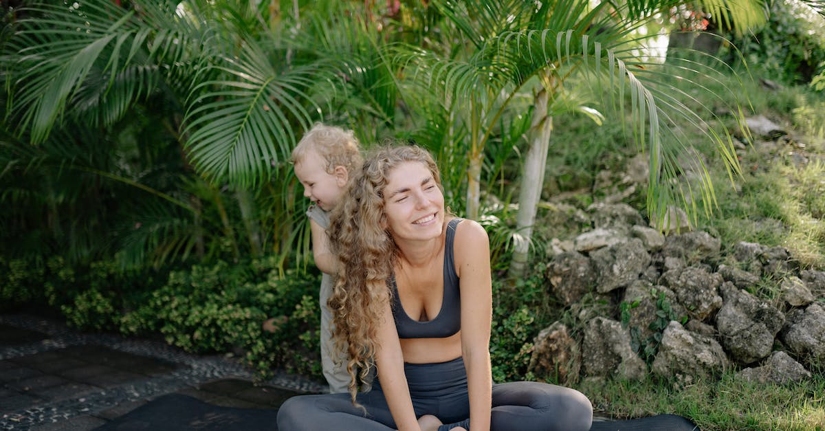 How do I play well as a scout? - Cheerful smiling sportswoman sitting in lotus pose with adorable little son on yoga mat in green tropical garden