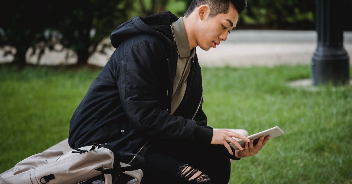 How do I use survival trail scrolls? - Side view of focused young Asian male student in casual clothes sitting on bench in park with backpack and browsing internet on tablet