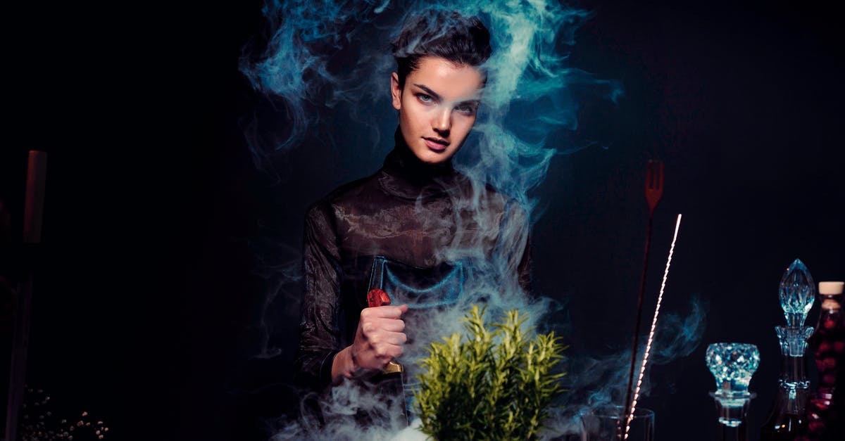How do you catch a fairy in a bottle - Graceful young female alchemist with knife in hand in black outfit preparing potion from various herbs among smoke in dark room