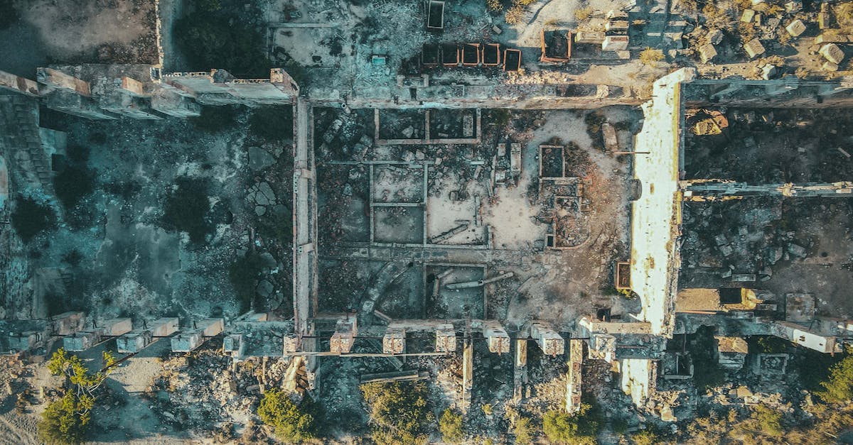 How do you view your town's history when sitting on the wall in the event plaza? - Drone view of ruined stone buildings with damaged walls and roofs located in uninhabited district