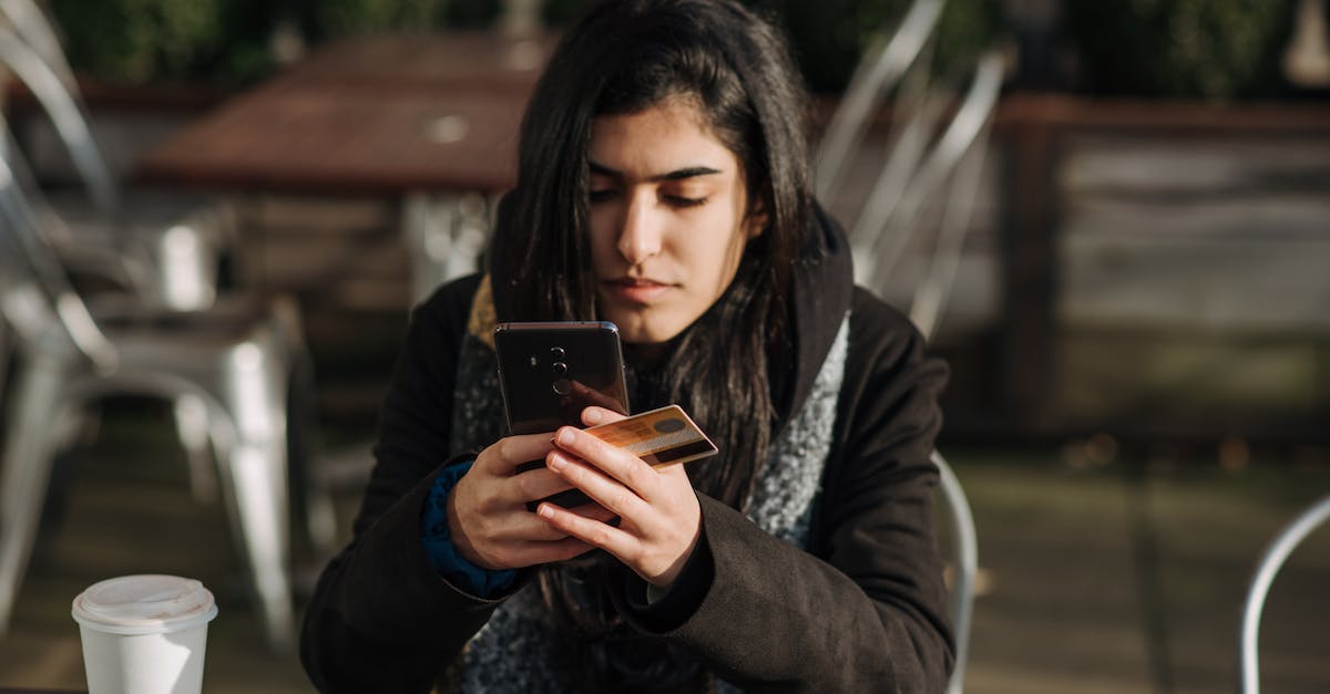 How to buy free game? - Young ethnic female purchaser with credit card shopping online on mobile phone at table with takeaway hot drink in cafeteria