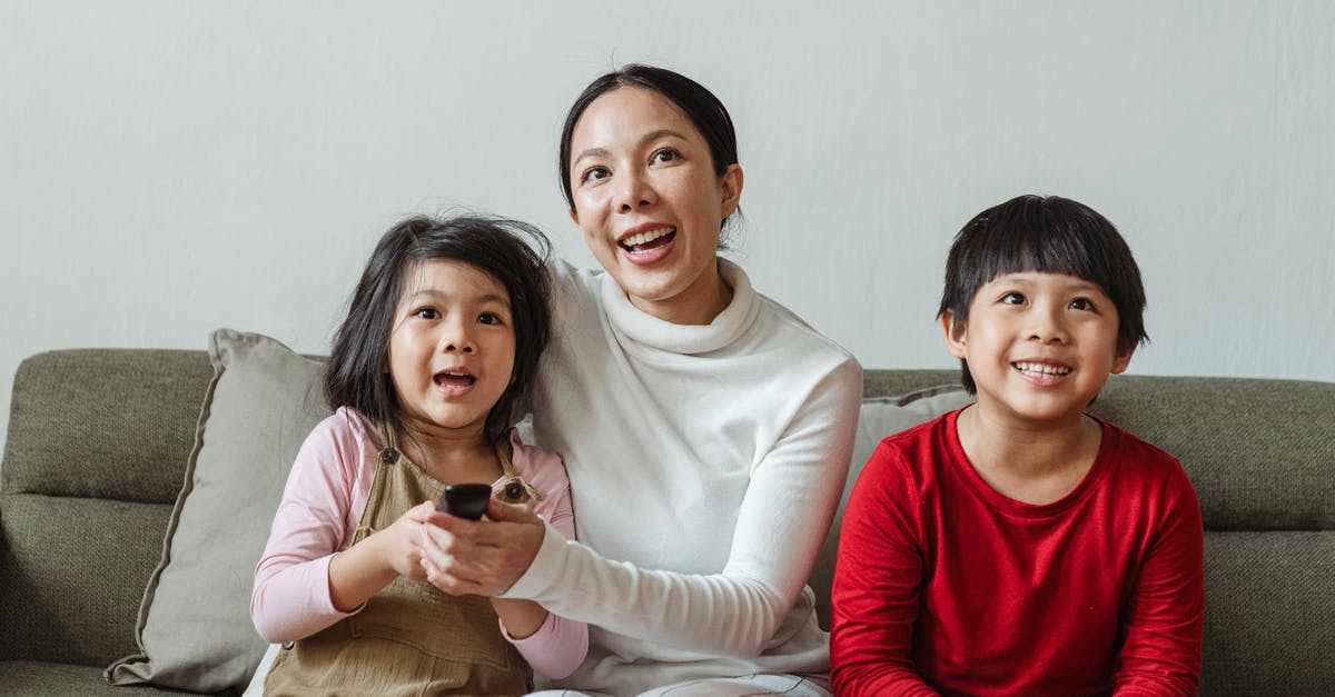 How to choose save device for downloads on Xbox Live - Optimistic Asian woman with little daughter and son in casual outfit sitting on sofa while choosing television channel using remote