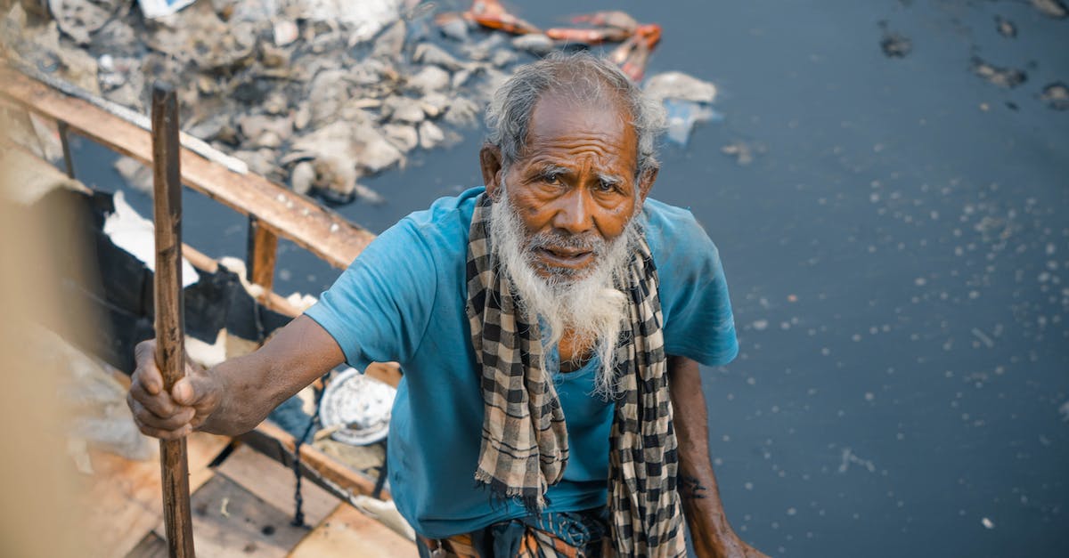 How to cope with local domination? - From above cheerless elderly Asian male in worn out clothes standing near dirty polluted river full of rubbish in poor district and looking at camera