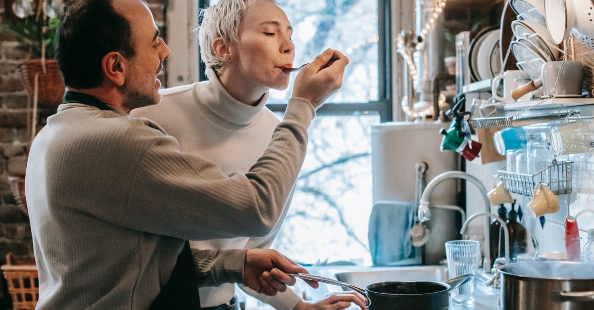 How to feed hopper when owned by prisoners? - Side view of content ethnic man in casual clothes and apron feeding happy wife while preparing delicious dinner at stove in kitchen