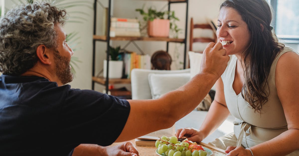How to give wheat to a hungry follower? - Positive adult man giving fresh grape to wife while sitting together at wicker table with various fruits in cozy apartment in daylight