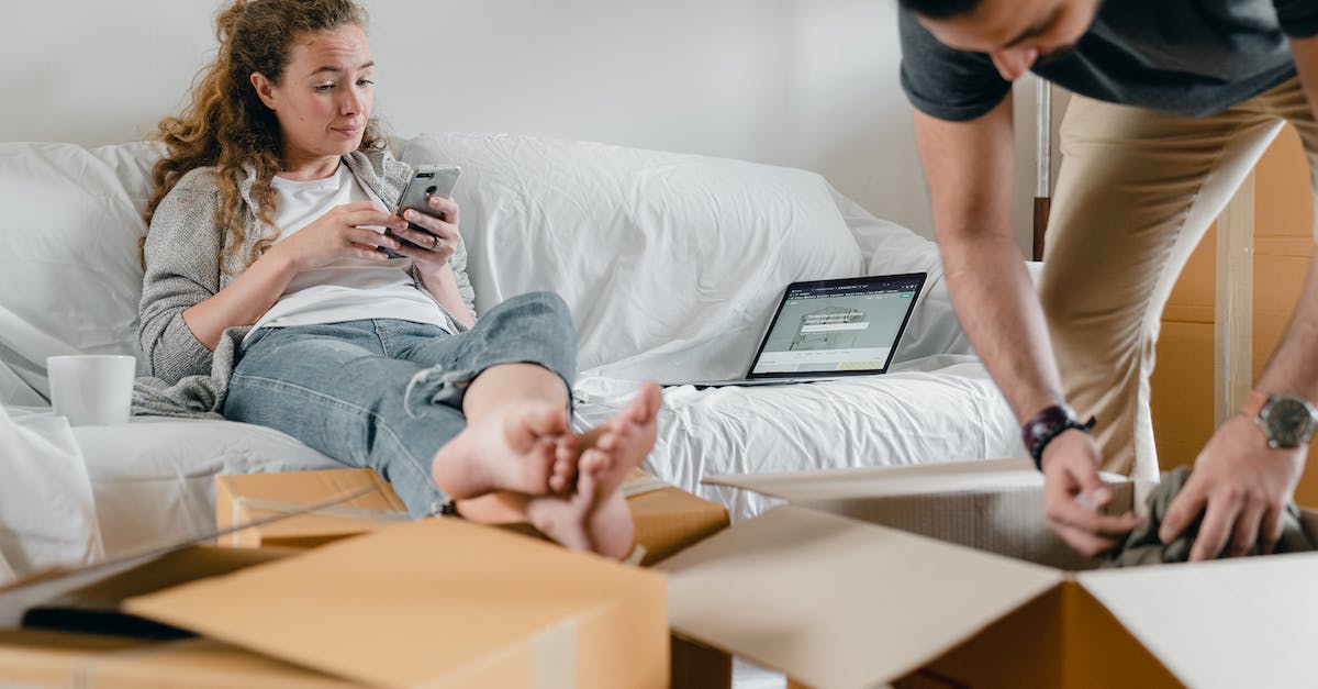 How to insert emoticons in the chat box? - Barefoot female in casual wear sitting with crossed legs on sofa with open laptop while using social media on cellphone and crop male partner getting clothes out of cardboard box